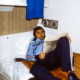 Young man in a blue tracksuit lounging on a bed, reading a newspaper in a small, retro-style room.