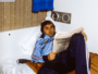 Young man in a blue tracksuit lounging on a bed, reading a newspaper in a small, retro-style room.