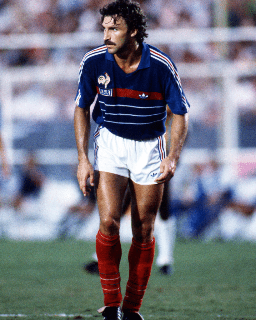 Soccer player in a blue jersey with red stripe and white shorts, standing on a grass field during a match with red socks visible.