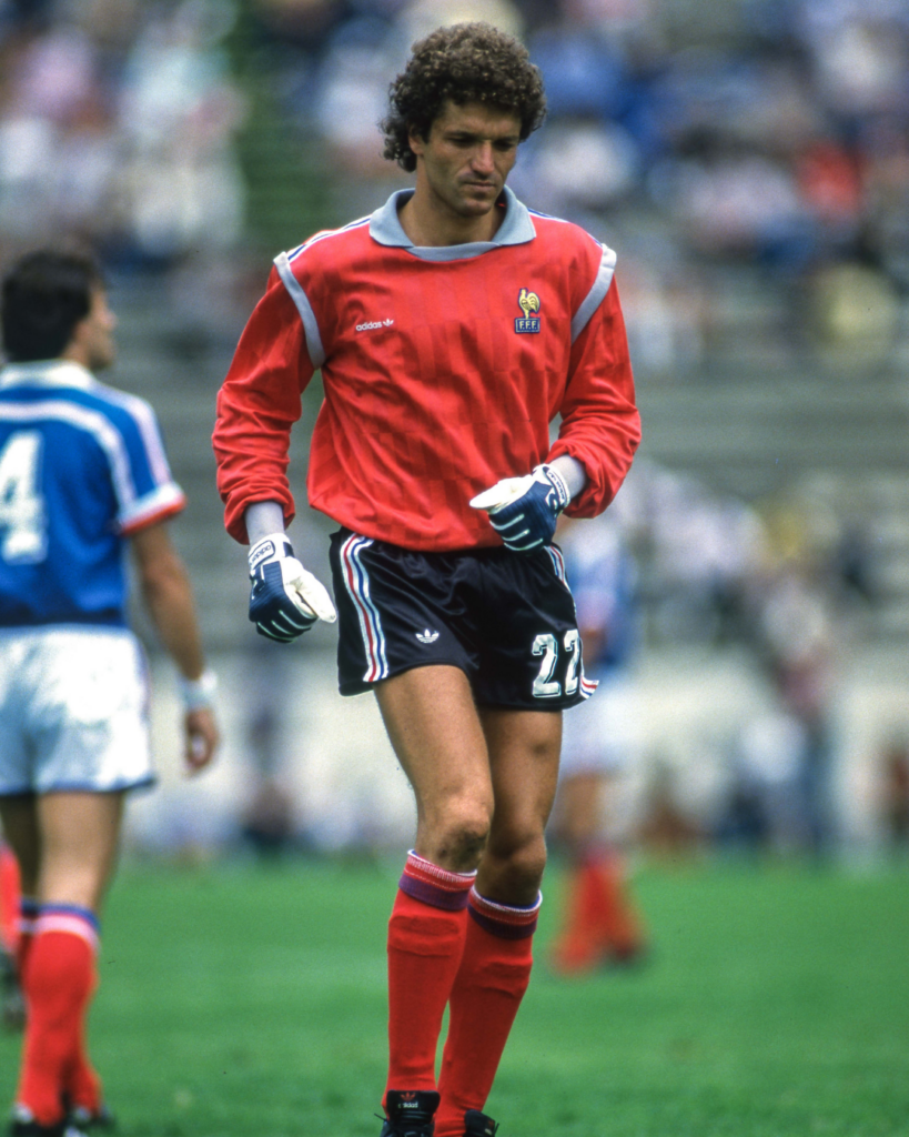 Soccer goalkeeper in a red jersey and blue shorts standing on a grassy field, wearing gloves.