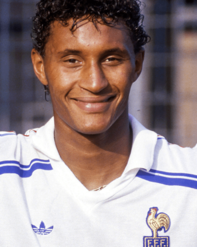 Close-up of a smiling man wearing a white soccer jersey with blue stripes and a French Football Federation crest outdoors.