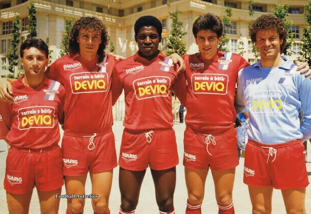 645685141_4371413896470928_5281499083635643353_n | AllezPaillade.com Five male soccer players stand arm in arm outdoors, wearing red uniforms with a yellow 'DEVIQ' sponsor logo and white drawstrings; a blue-jersey goalkeeper stands on the right.
