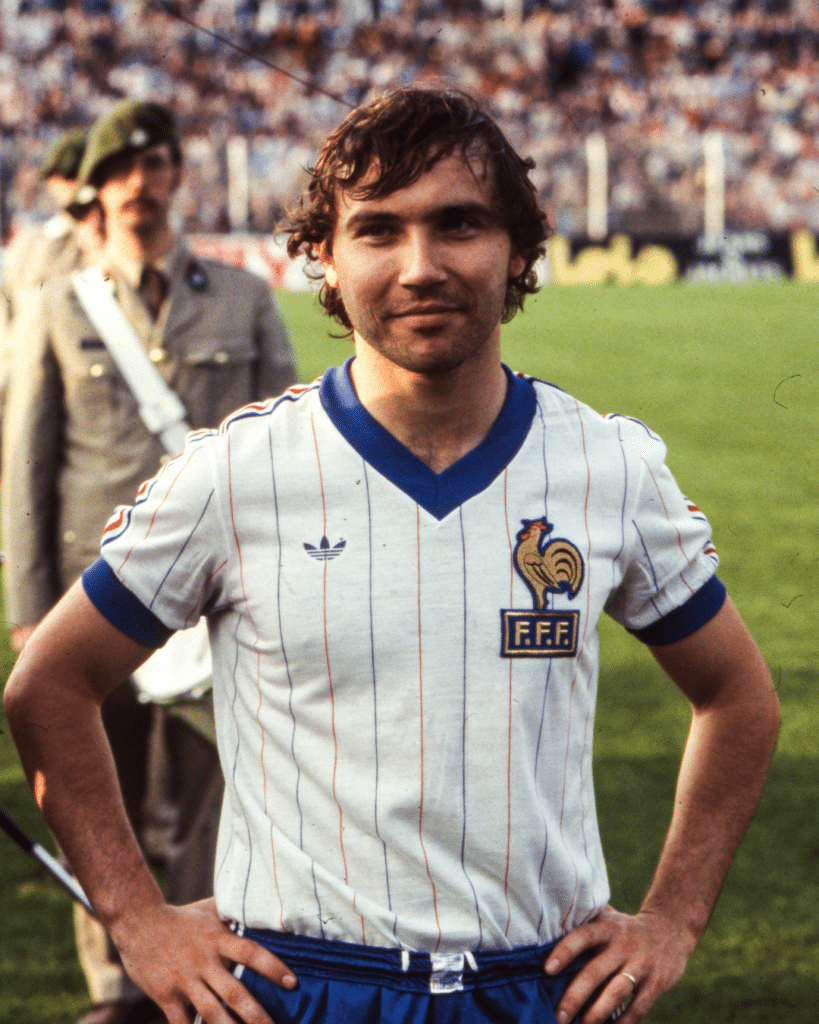 French footballer in a white pinstriped France kit with blue collar and rooster crest, standing with hands on hips on a grass field.