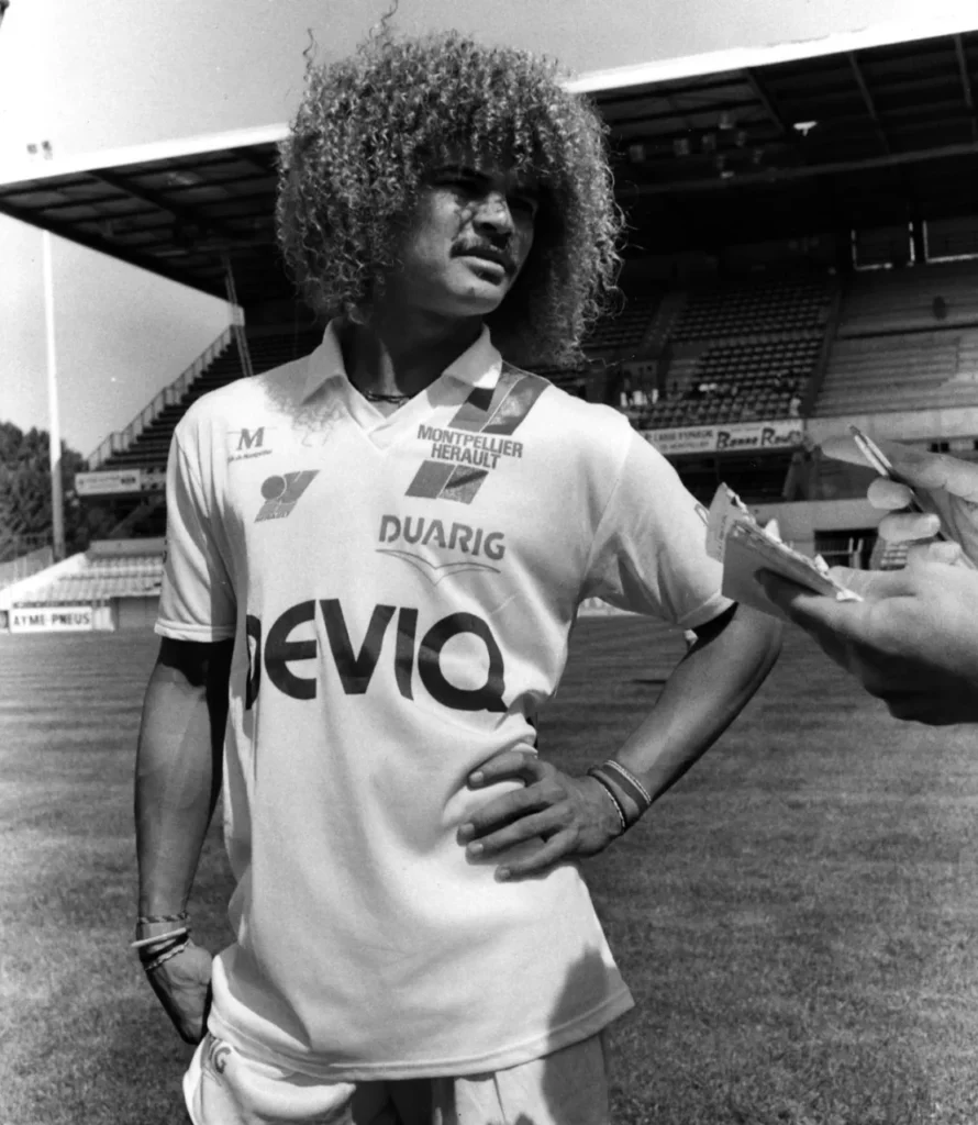 A-AAAAhhhh-quel-playboy-samuse-oulou-aime-tous-mes-joueurs-maislui-il-tait-spcial-est-marrant-il-a-toujours-la-mme-coupe-de-cheveux-aujourdhui-sais | AllezPaillade.com Black-and-white photo of a footballer with a large curly hairstyle being interviewed on a sunlit stadium pitch, hands on hips, reporters' hands with notebooks nearby.