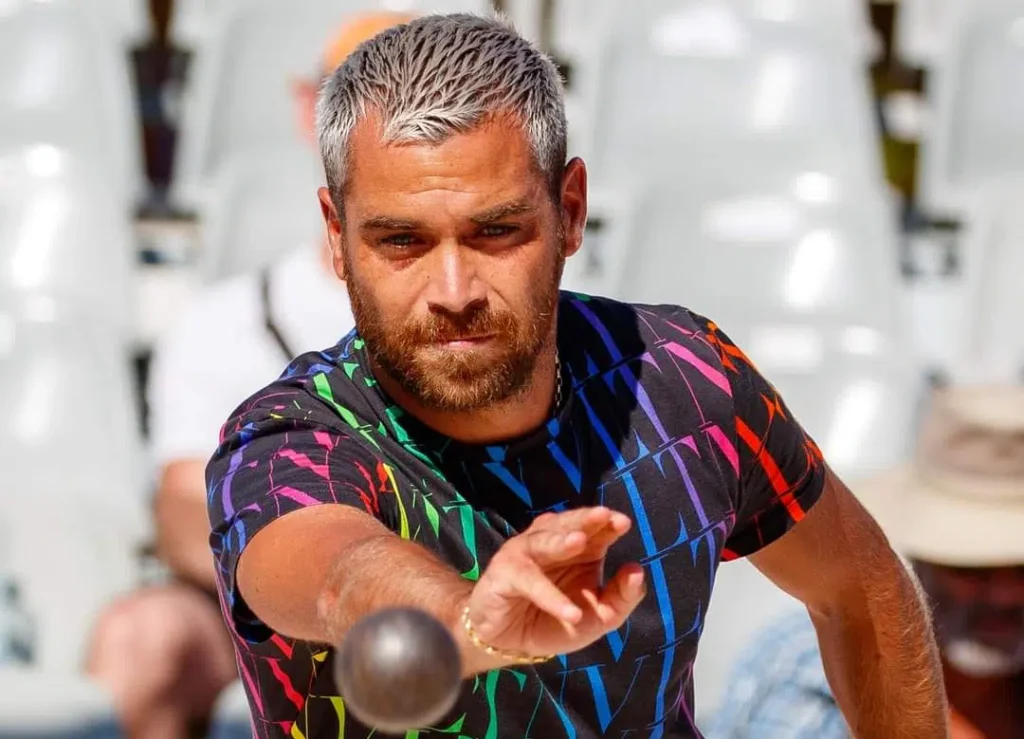 Man with short gray hair and beard in a colorful patterned shirt releasing a bowling ball towards the pins in a bowling alley.