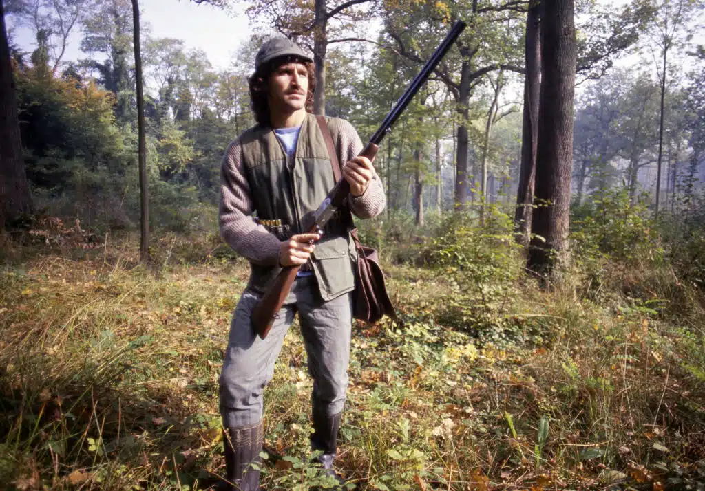 Daniel Xuereb | AllezPaillade.com Hunter standing in a forest clearing, wearing a vest and hat, holding a rifle diagonally across his body.