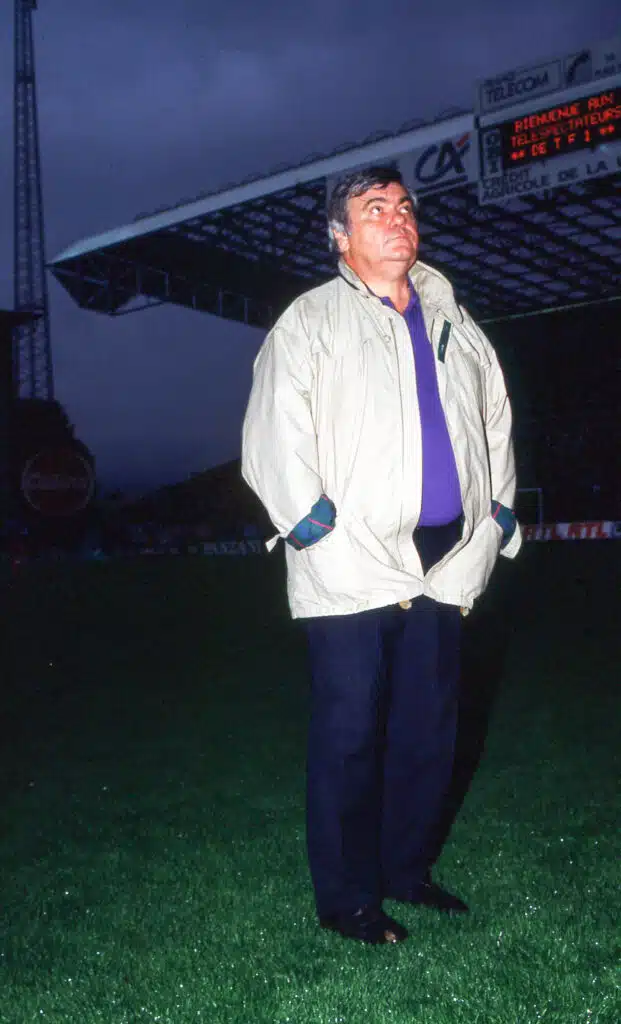 Saint Etienne v Montpellier - Division 1 | AllezPaillade.com Man standing on a grassy stadium field at dusk, wearing a beige jacket and purple shirt, hands in pockets.
