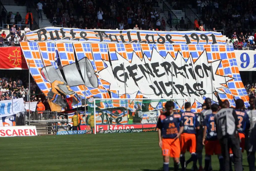 Supporters de Montpellier | AllezPaillade.com Soccer stadium scene with players on the field and a large colorful banner in the stands above the goal, surrounded by crowds of spectators in the seats.
