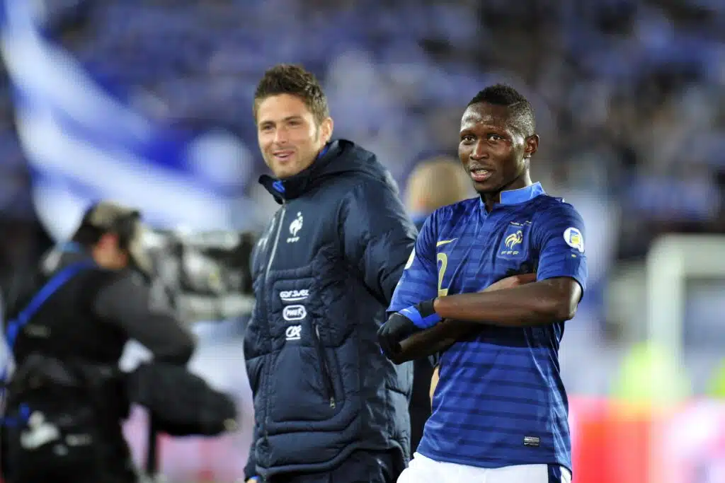Olivier GIROUD Mapou YANGAMBIWA | AllezPaillade.com Two male soccer players stand side by side on the pitch, one in a blue striped France jersey and the other in a dark training jacket, smiling at the camera.