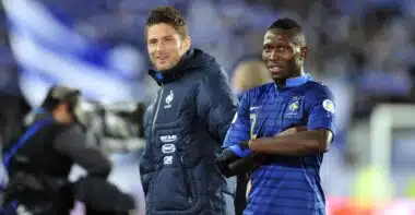 Olivier GIROUD Mapou YANGAMBIWA | AllezPaillade.com Two male soccer players stand side by side on the pitch, one in a blue striped France jersey and the other in a dark training jacket, smiling at the camera.