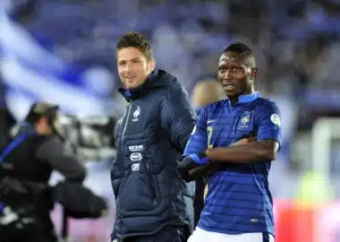 Olivier GIROUD Mapou YANGAMBIWA | AllezPaillade.com Two male soccer players stand side by side on the pitch, one in a blue striped France jersey and the other in a dark training jacket, smiling at the camera.