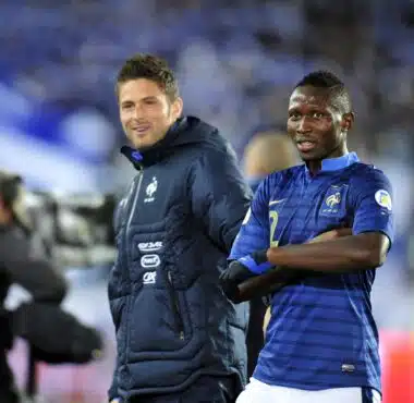 Two male soccer players stand side by side on the pitch, one in a blue striped France jersey and the other in a dark training jacket, smiling at the camera.