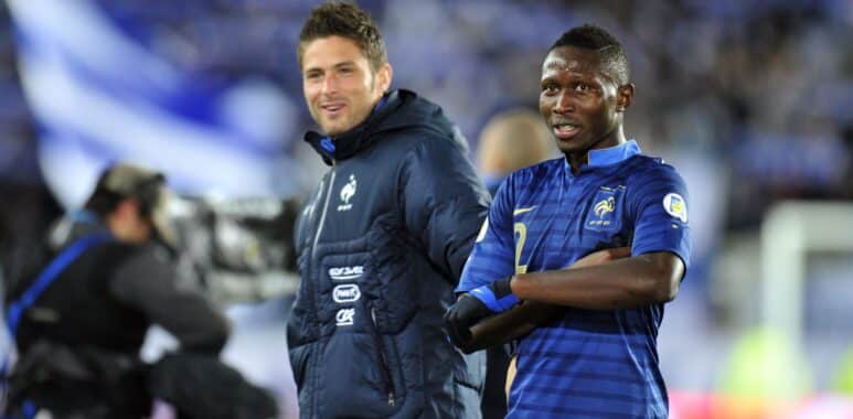 Olivier GIROUD Mapou YANGAMBIWA | AllezPaillade.com Two male soccer players stand side by side on the pitch, one in a blue striped France jersey and the other in a dark training jacket, smiling at the camera.