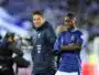 Two male soccer players stand side by side on the pitch, one in a blue striped France jersey and the other in a dark training jacket, smiling at the camera.