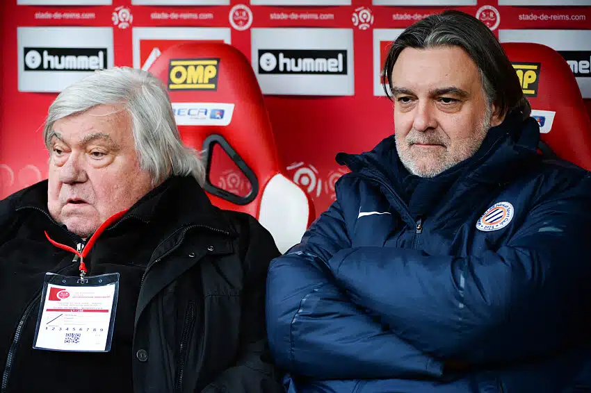 Louis Laurent Nicollin | AllezPaillade.com Two men sit side by side in a stadium dugout, wearing dark jackets, with red sponsor banners behind them.