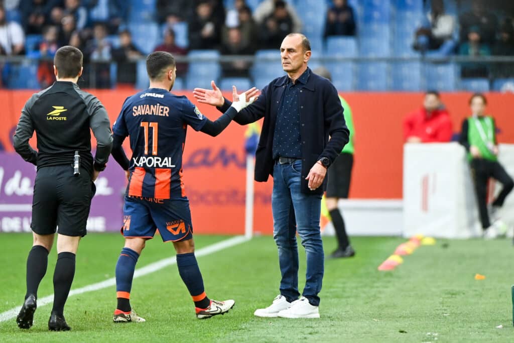 Soccer coach in a dark jacket shaking hands with a player in a blue and orange kit on the sideline during a match.