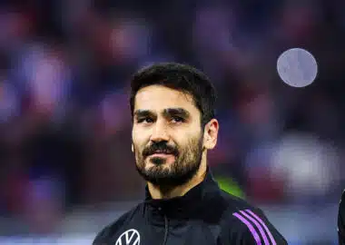 Portrait of a man with dark hair and a beard in a black jacket with purple stripes, standing in a sports arena.