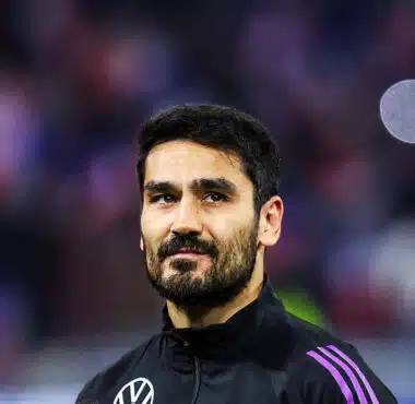 Portrait of a man with dark hair and a beard in a black jacket with purple stripes, standing in a sports arena.