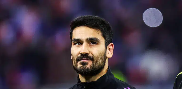 Gundogan | AllezPaillade.com Portrait of a man with dark hair and a beard in a black jacket with purple stripes, standing in a sports arena.