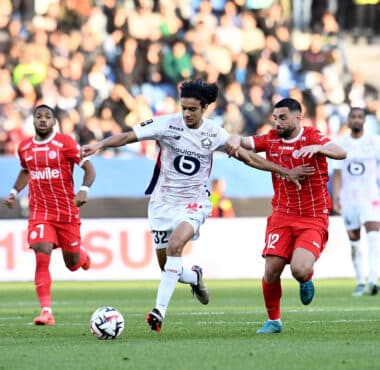 Soccer players in red and white battle for a ball on a green field with a blurred crowd in the stands behind them.
