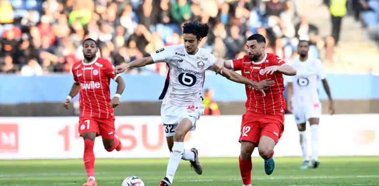 Soccer players in red and white battle for a ball on a green field with a blurred crowd in the stands behind them.