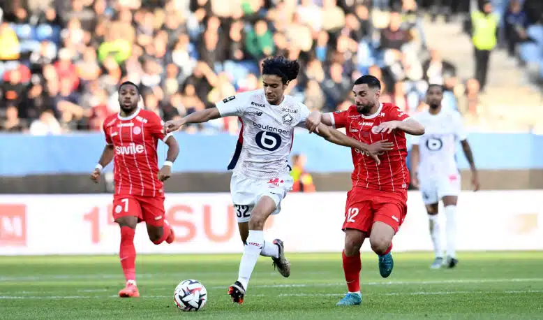 Soccer players in red and white battle for a ball on a green field with a blurred crowd in the stands behind them.