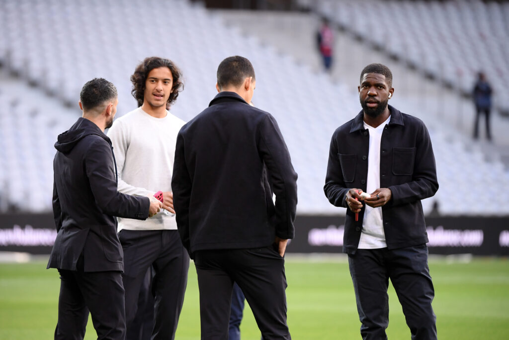 Four men in jackets stand together on a grass field, talking with blurred stadium seats in the background, suggesting a team discussion.