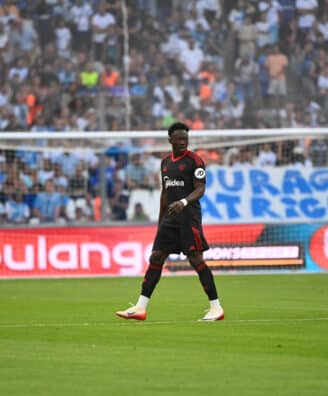 Soccer match scene with a Black player in a black kit with red trim in the foreground, crowd in the stands behind the goal.