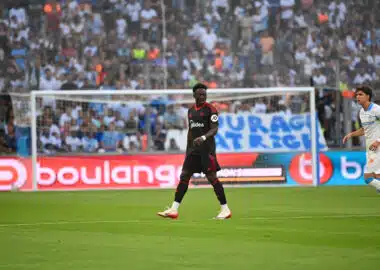 Soccer match scene with a Black player in a black kit with red trim in the foreground, crowd in the stands behind the goal.