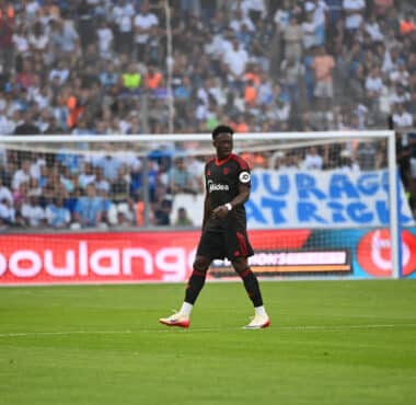Soccer match scene with a Black player in a black kit with red trim in the foreground, crowd in the stands behind the goal.