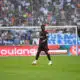 Soccer match scene with a Black player in a black kit with red trim in the foreground, crowd in the stands behind the goal.