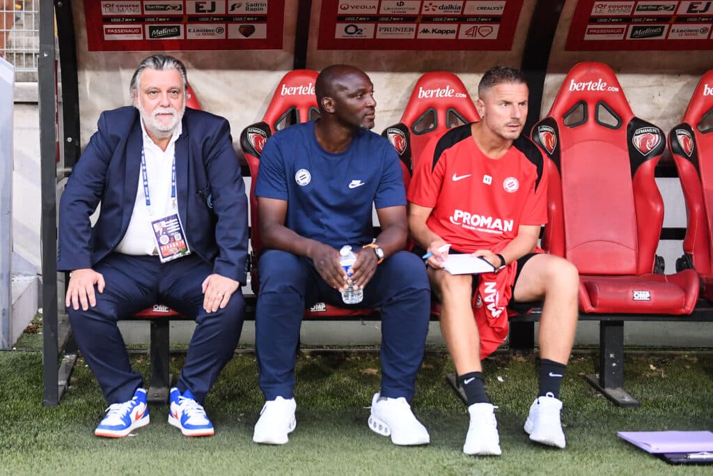 Three men sit on a dugout bench: an older man in a suit on the left, a man in blue in the middle, and a player in a red kit on the right.