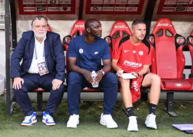 Le Mans FC v Montpellier Herault SC - Ligue 2 BKT 20252026 | AllezPaillade.com Three men sit on a dugout bench: an older man in a suit on the left, a man in blue in the middle, and a player in a red kit on the right.