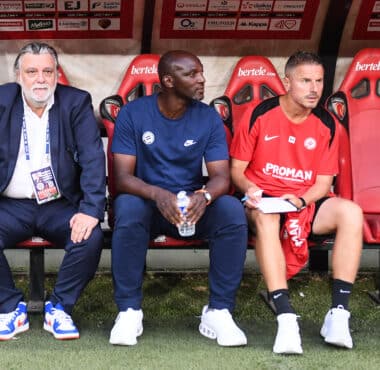 Three men sit on a dugout bench: an older man in a suit on the left, a man in blue in the middle, and a player in a red kit on the right.