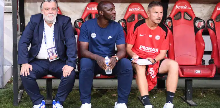 Three men sit on a dugout bench: an older man in a suit on the left, a man in blue in the middle, and a player in a red kit on the right.