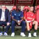 Three men sit on a dugout bench: an older man in a suit on the left, a man in blue in the middle, and a player in a red kit on the right.