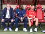Three men sit on a dugout bench: an older man in a suit on the left, a man in blue in the middle, and a player in a red kit on the right.
