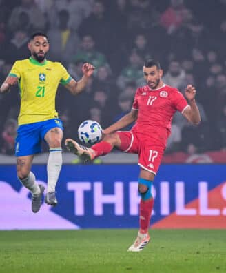 Skhiri Brazil v Tunisia - Friendly Match 18112025 | AllezPaillade.com Two soccer players—Brazil in yellow, Tunisia in red—jump for the ball during a match, crowd in the stands behind them.