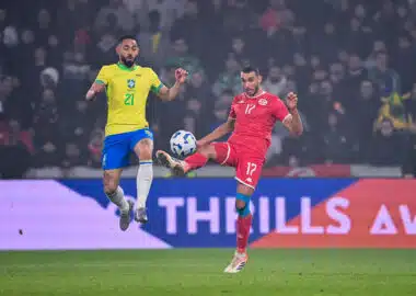 Two soccer players&mdash;Brazil in yellow, Tunisia in red&mdash;jump for the ball during a match, crowd in the stands behind them.