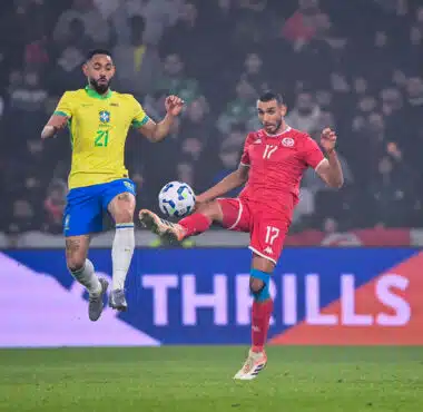 Two soccer players&mdash;Brazil in yellow, Tunisia in red&mdash;jump for the ball during a match, crowd in the stands behind them.