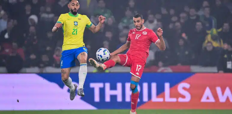 Two soccer players&mdash;Brazil in yellow, Tunisia in red&mdash;jump for the ball during a match, crowd in the stands behind them.