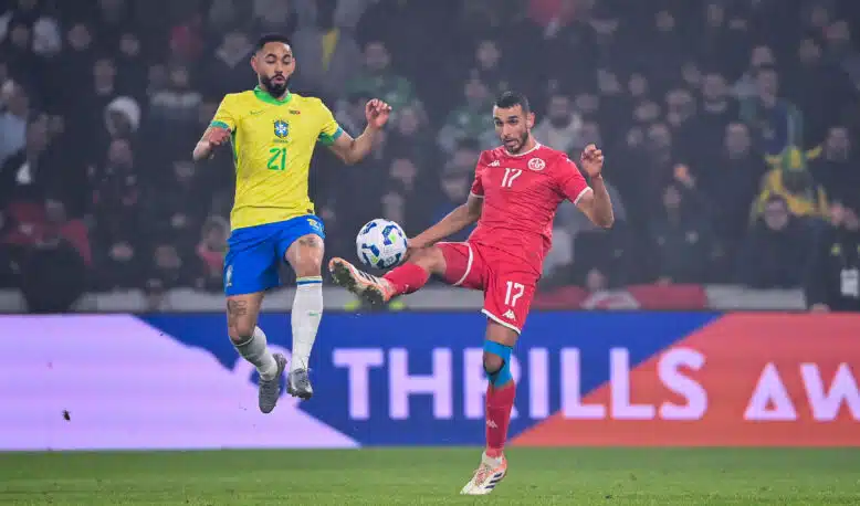 Two soccer players&mdash;Brazil in yellow, Tunisia in red&mdash;jump for the ball during a match, crowd in the stands behind them.