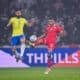 Two soccer players&mdash;Brazil in yellow, Tunisia in red&mdash;jump for the ball during a match, crowd in the stands behind them.