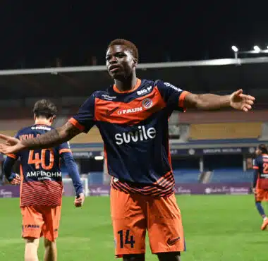 Soccer player in orange and navy kit celebrates with arms outstretched on a stadium field at night.