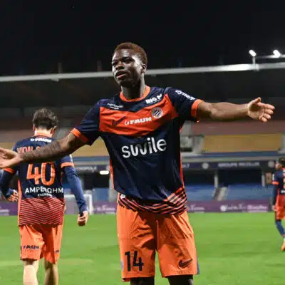 Soccer player in orange and navy kit celebrates with arms outstretched on a stadium field at night.