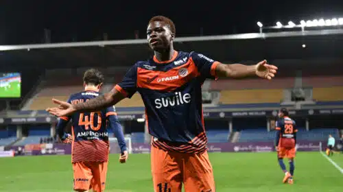 Soccer player in orange and navy kit celebrates with arms outstretched on a stadium field at night.