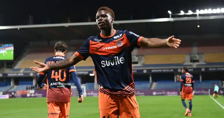Soccer player in orange and navy kit celebrates with arms outstretched on a stadium field at night.