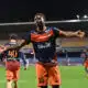 Soccer player in orange and navy kit celebrates with arms outstretched on a stadium field at night.