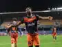 Soccer player in orange and navy kit celebrates with arms outstretched on a stadium field at night.