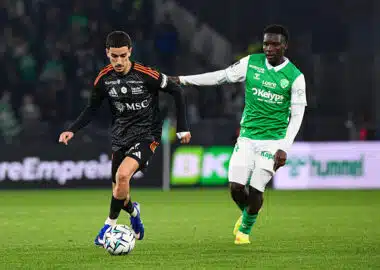 Soccer players contest the ball on a green field; one in black with orange accents, the other in green and white.